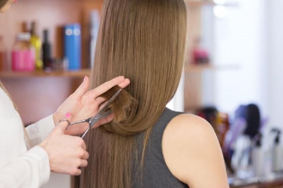 Người thợ cắt tóc đang tỉa tóc dài của khách hàng trong một tiệm làm tóc, với các sản phẩm chăm sóc tóc phía sau.