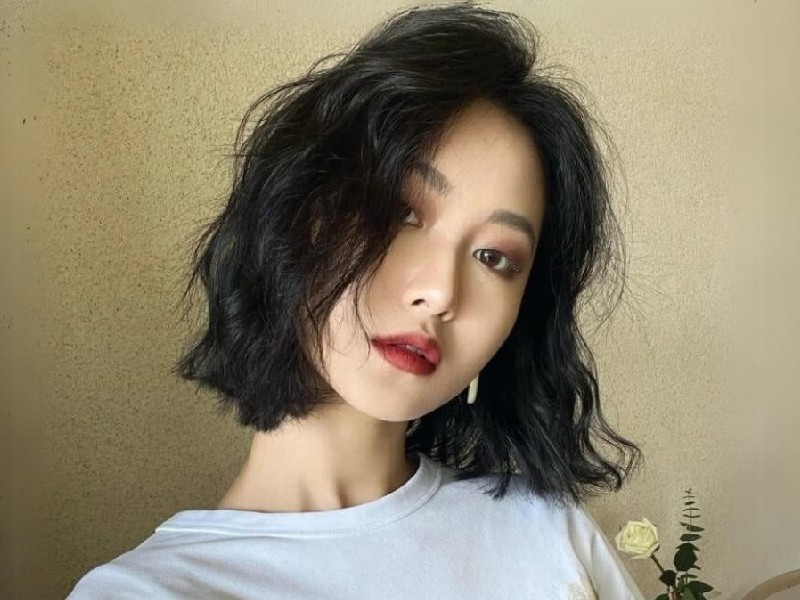 Người với tóc ngắn đen, mặc áo trắng, đứng trước nền tường màu be, biểu cảm tự nhiên.