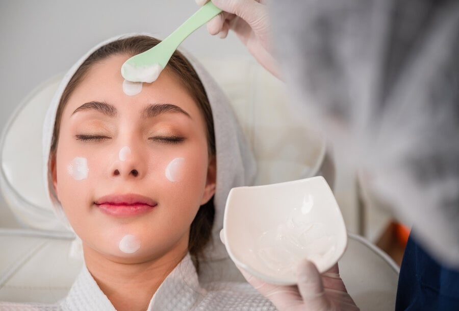 Người đang thư giãn trong spa, được thoa kem dưỡng lên mặt với thìa nhựa xanh nhạt.