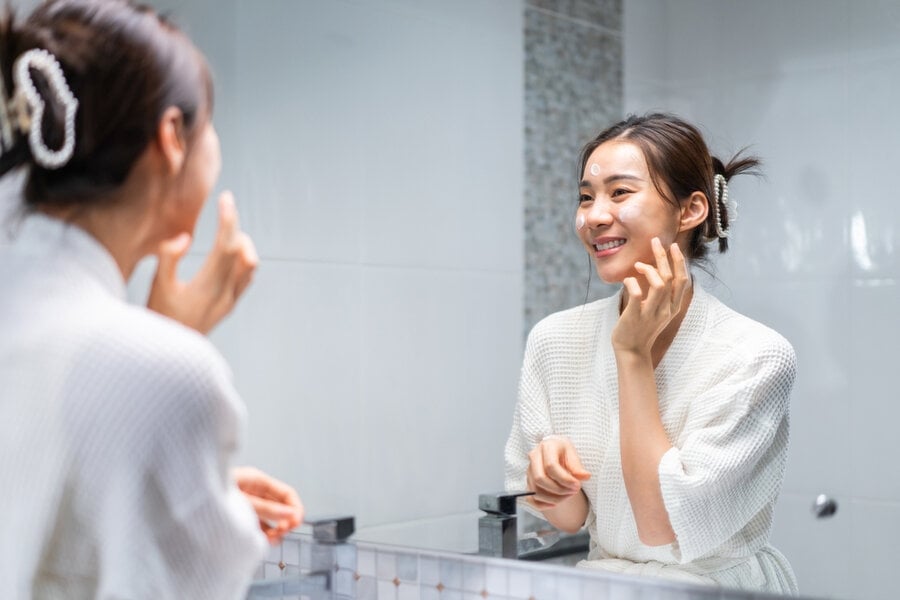 Người phụ nữ mặc áo choàng tắm, đứng trước gương, bôi sản phẩm chăm sóc da lên mặt và mỉm cười.
