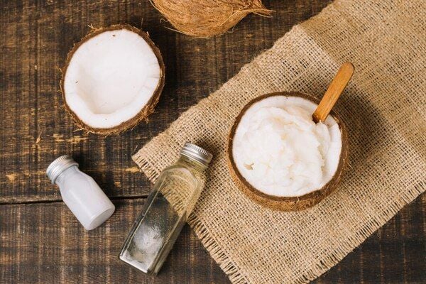Nửa quả dừa, dầu dừa trong chai thủy tinh và dừa nạo trong bát gỗ trên nền vải bố và bàn gỗ.