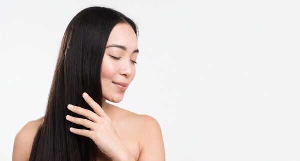 Người phụ nữ với mái tóc dài mượt, tay chạm nhẹ vào tóc, biểu cảm thư giãn, nền trắng.