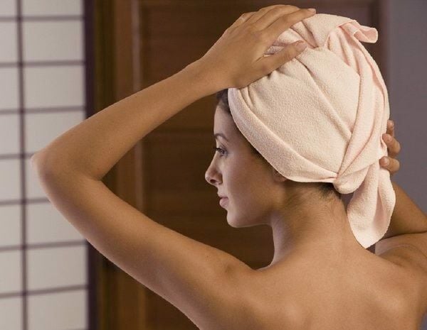 boc_keratin_truc_tiep_cho_toc_ad01fb89c36945468c5e8b0c12c0c74c_grande Người phụ nữ quấn khăn tắm trên đầu, đứng trong phòng có ánh sáng nhẹ.