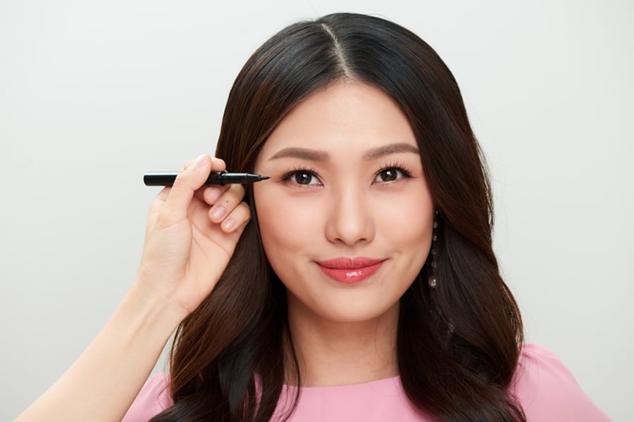 Người phụ nữ đang được kẻ mắt bằng bút kẻ mắt, mặc áo hồng và có mái tóc đen dài.