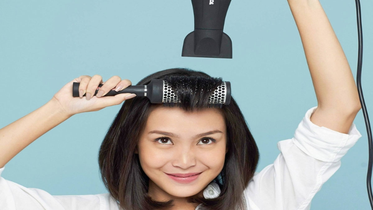 cach-cat-toc-layer-tai-nha-6 Người phụ nữ cầm máy sấy tóc và lược tròn, đang tạo kiểu tóc trên nền xanh dương nhạt.