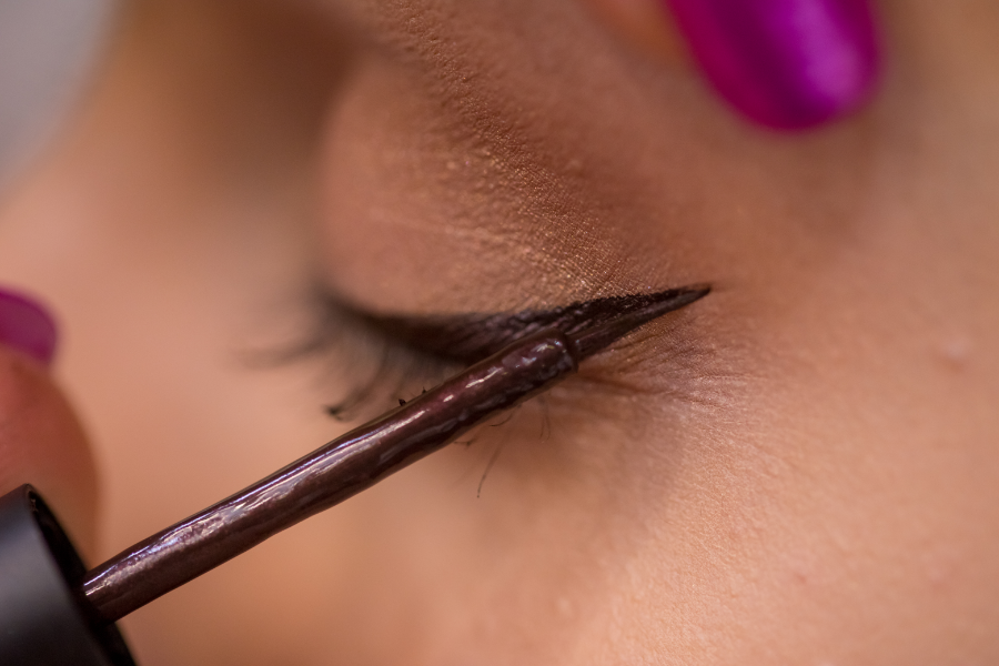 Cận cảnh một người đang vẽ eyeliner màu nâu lên mí mắt, nền mắt có màu nâu nhạt, móng tay màu tím.