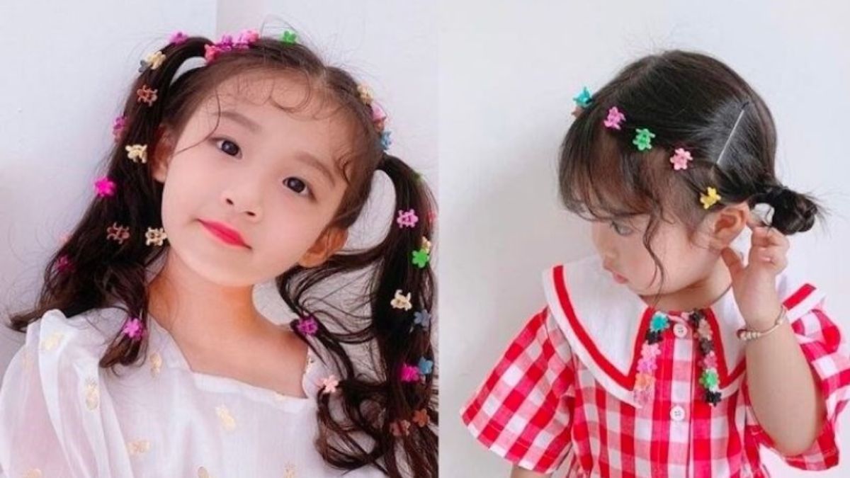 Hai bé gái với tóc buộc, trang trí bằng kẹp hoa nhiều màu sắc, mặc áo trắng và áo kẻ caro đỏ.