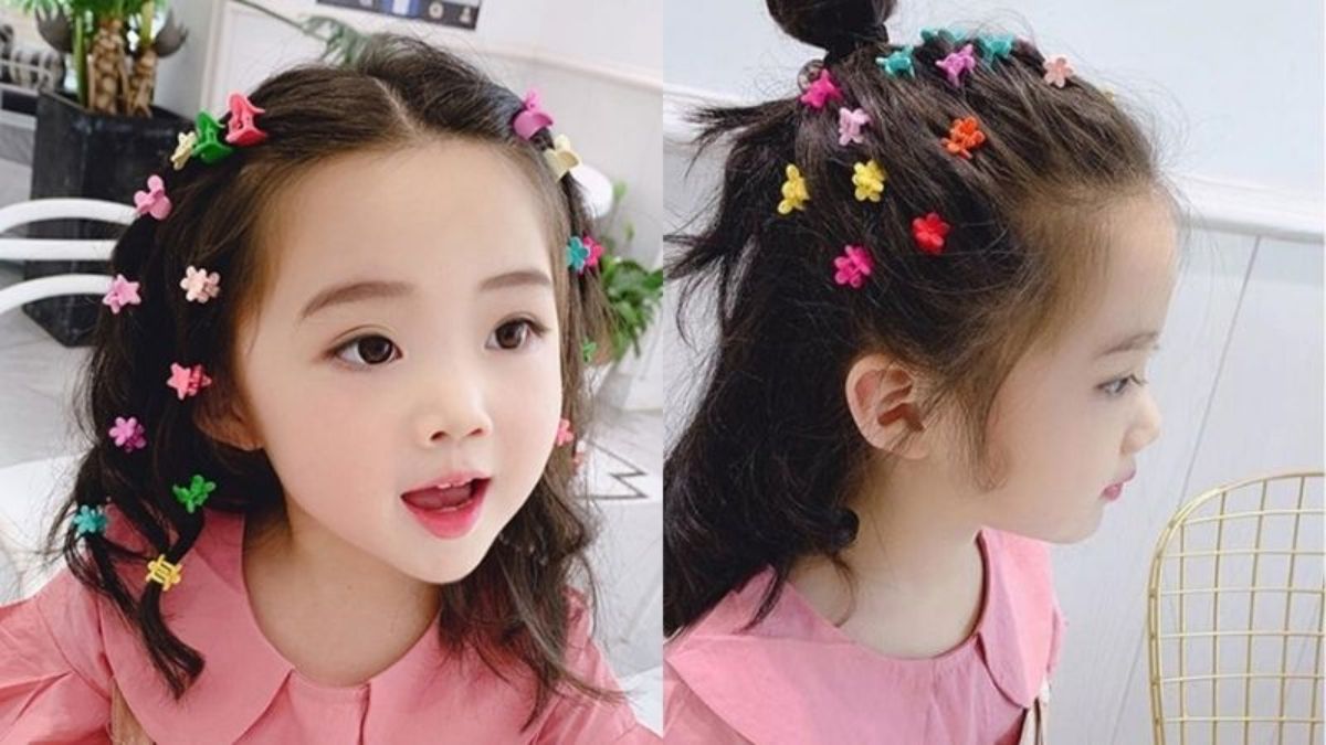 Bé gái với tóc cài nhiều kẹp hoa màu sắc, mặc áo hồng, trong không gian nội thất sáng sủa.