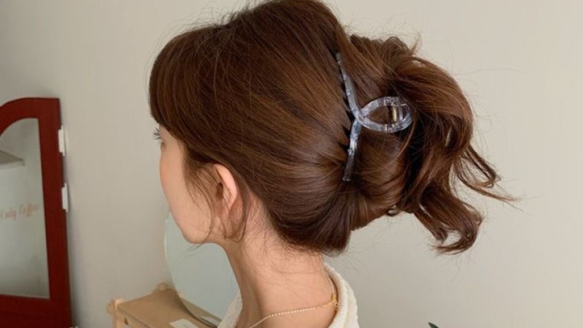 Người đang búi tóc nâu bằng kẹp tóc lớn, đứng trước gương trong phòng có tông màu ấm.