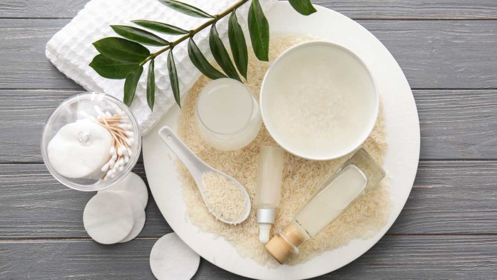 cach-lam-toc-muot-3 Bộ sưu tập sản phẩm chăm sóc da từ gạo, gồm bát gạo, chai lọ, bông tẩy trang và nhánh lá xanh trên nền gỗ xám.
