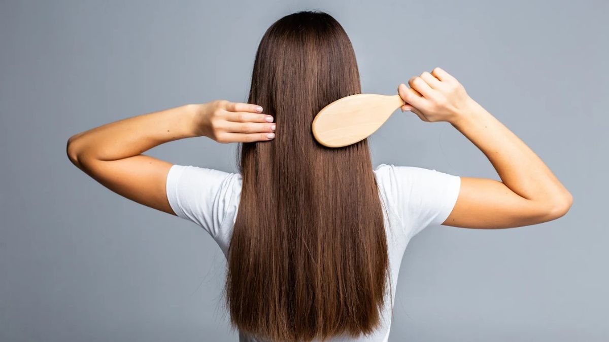 Người mặc áo trắng đang chải tóc dài màu nâu bằng lược gỗ, chụp từ phía sau.
