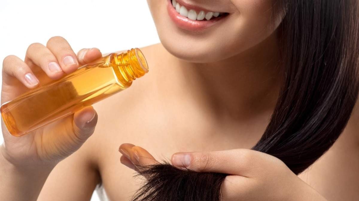 Người cầm chai dầu dưỡng màu vàng, áp dụng lên tóc đen bóng, gương mặt tươi cười phía sau.