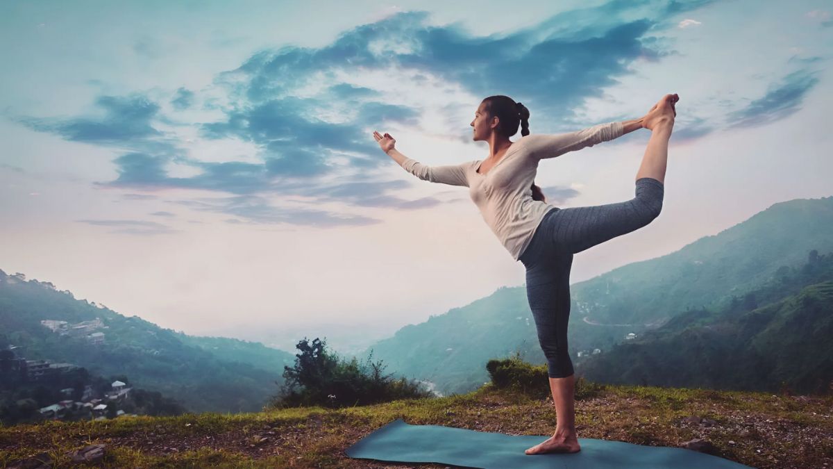 Người tập yoga trong tư thế vũ công trên thảm, khung cảnh núi non và bầu trời xanh phía sau.
