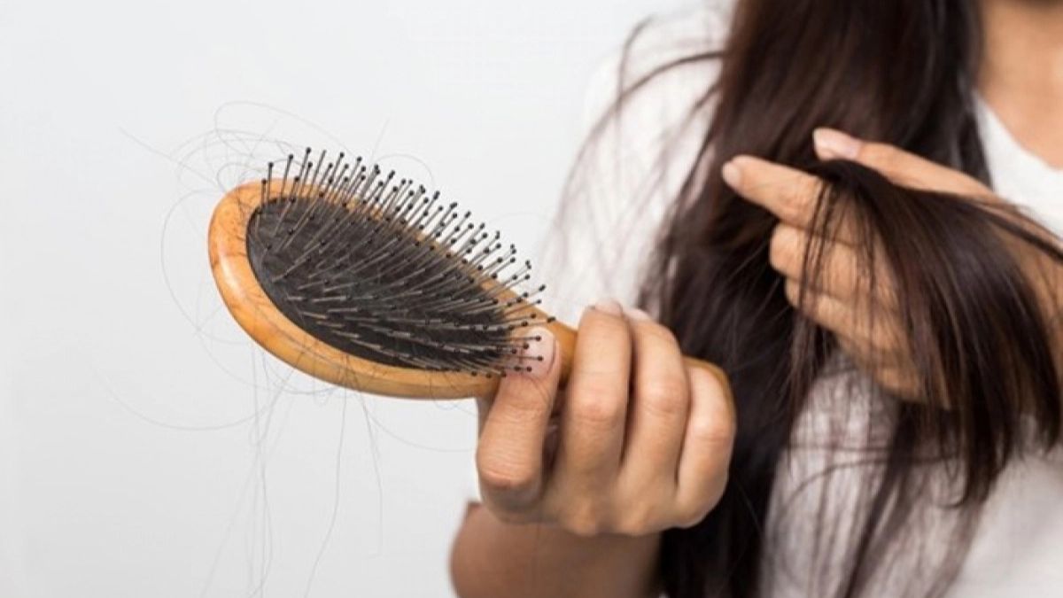 Người cầm bàn chải tóc với nhiều sợi tóc rụng, tay kia cầm một lọn tóc dài, nền trắng.