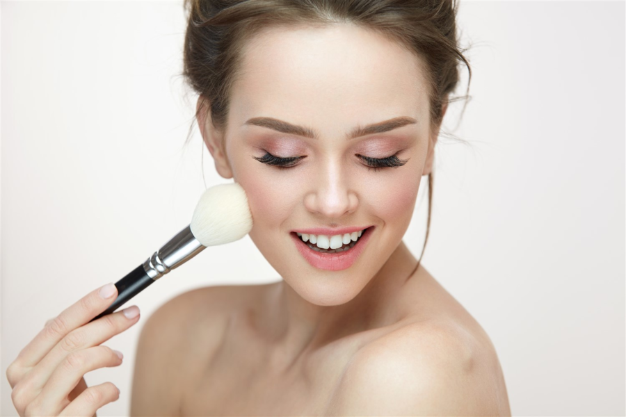 Người cầm cọ trang điểm, đang mỉm cười và áp cọ lên má, nền sáng.