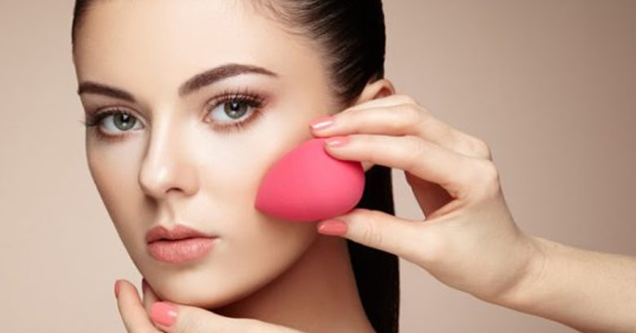Người cầm mút trang điểm màu hồng áp lên má, nền màu be.