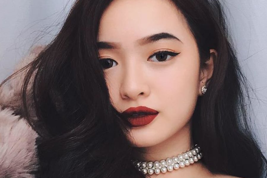 Người phụ nữ với trang điểm đậm, đeo vòng cổ ngọc trai, tóc dài, biểu cảm tự tin.
