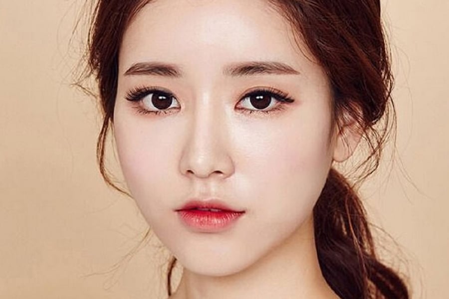 Chân dung một người với mái tóc nâu dài, trang điểm nhẹ, nhìn thẳng vào máy ảnh, nền màu be.