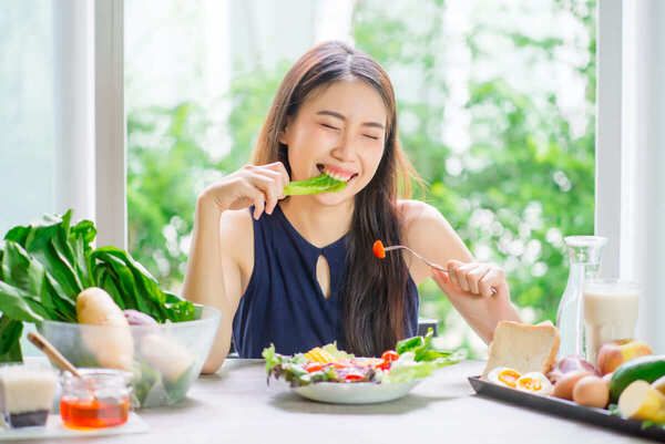 Người phụ nữ ngồi ăn salad rau củ tại bàn, xung quanh có nhiều thực phẩm tươi như rau, trái cây và bánh mì.