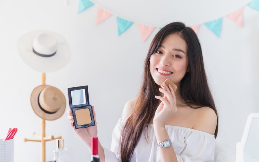 Người phụ nữ cầm hộp phấn trang điểm, mỉm cười, ngồi trước bàn với mũ và đồ trang điểm xung quanh.