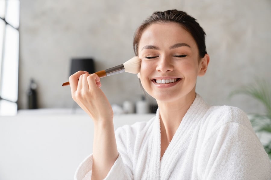 Người phụ nữ trong áo choàng tắm trắng, mỉm cười khi đang trang điểm với cọ trên má, nền phòng tắm mờ.