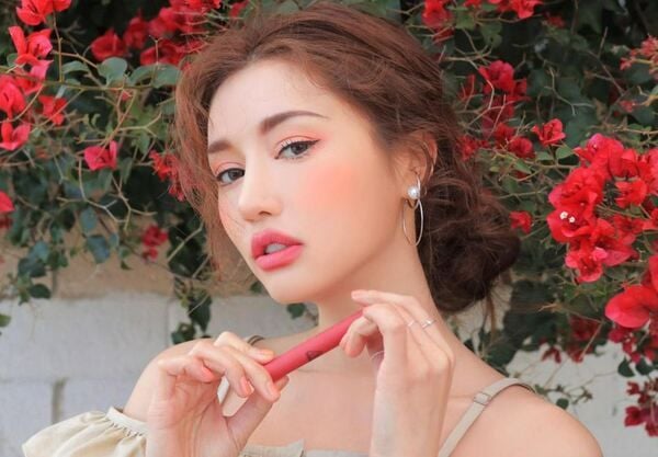 Người phụ nữ cầm son, đứng trước cây hoa đỏ, tóc búi, trang điểm tinh tế, ánh sáng tự nhiên.