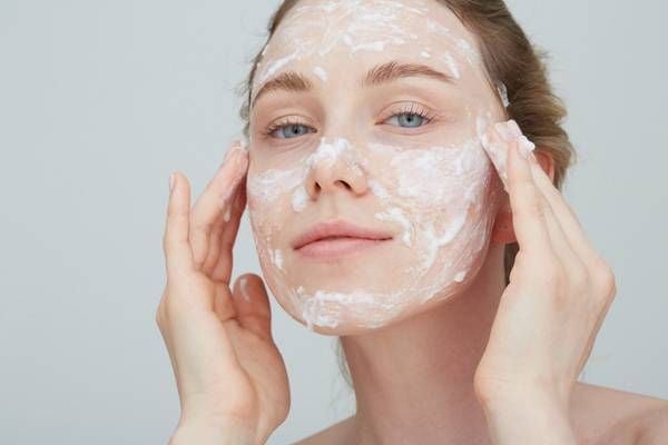 Người đang thoa mặt nạ dưỡng da, hai tay chạm nhẹ lên mặt, nền màu xám nhạt.