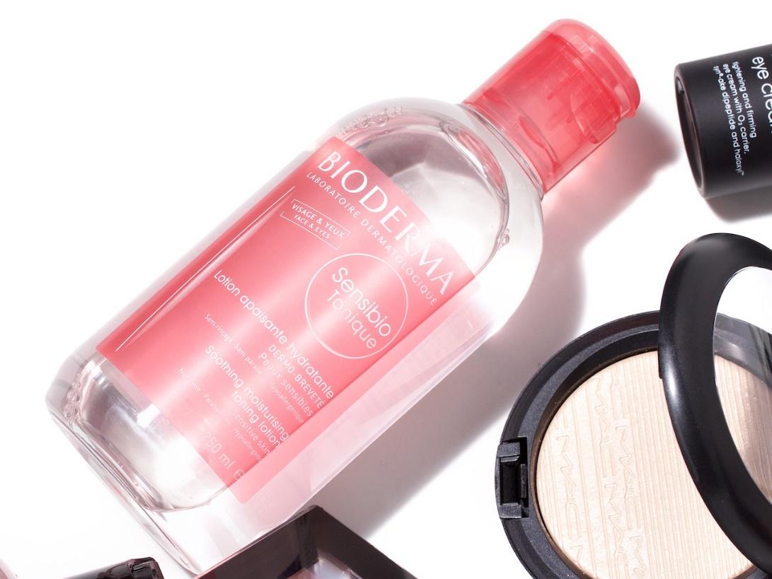 Chai nước tẩy trang c&oacute; nắp đỏ nằm cạnh một hộp phấn trang điểm v&agrave; một lọ mỹ phẩm nhỏ m&agrave;u đen.