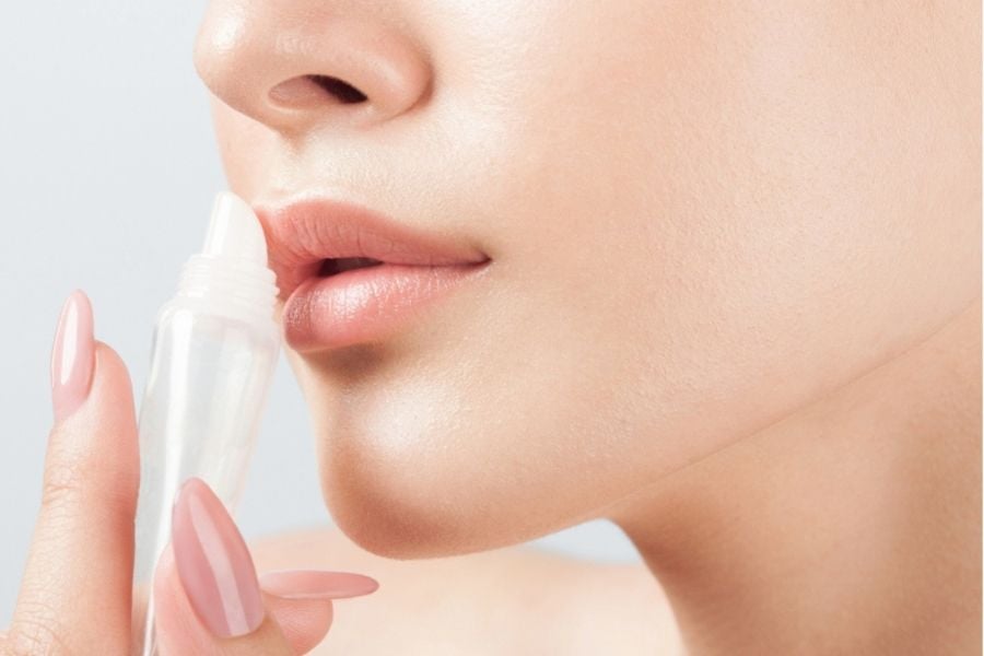 Người đang thoa son dưỡng lên môi từ một ống tuýp nhựa trong suốt, móng tay sơn màu hồng nhạt.