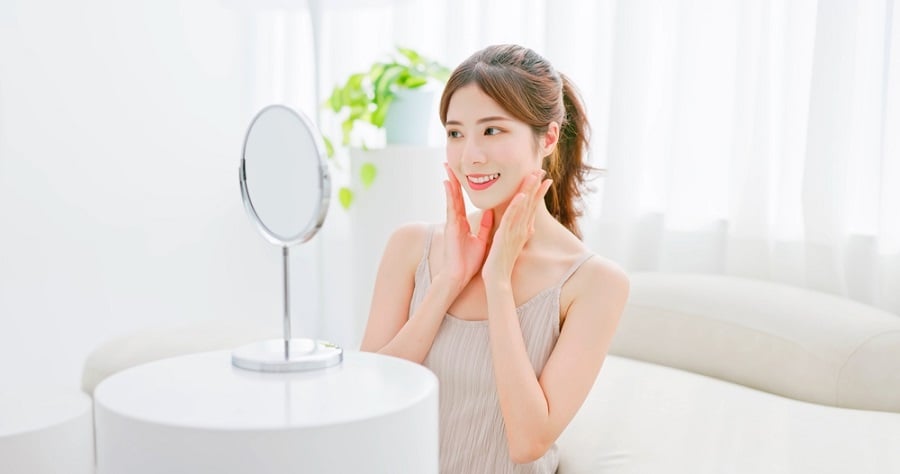 Người phụ nữ ngồi trước gương, mỉm cười và chạm vào mặt, trong không gian sáng sủa với cây xanh phía sau.