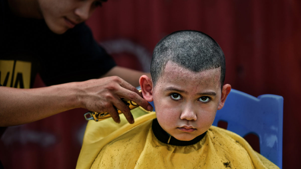 dau-nam-co-nen-cat-toc-5 Cậu bé ngồi trên ghế xanh, mặc áo choàng vàng, đang được cắt tóc bởi một người dùng tông đơ.