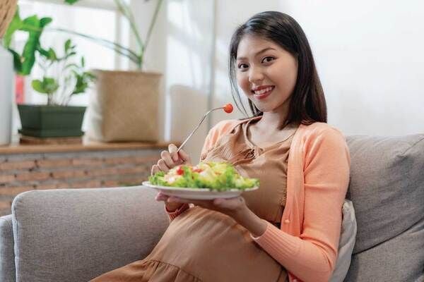 Người phụ nữ ngồi trên ghế sofa, cầm đĩa salad và nĩa, có cây xanh trang trí phía sau.