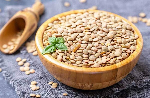 Bát gỗ chứa đầy đậu lăng khô, bên cạnh có một chiếc xẻng gỗ nhỏ trên nền vải màu xám.