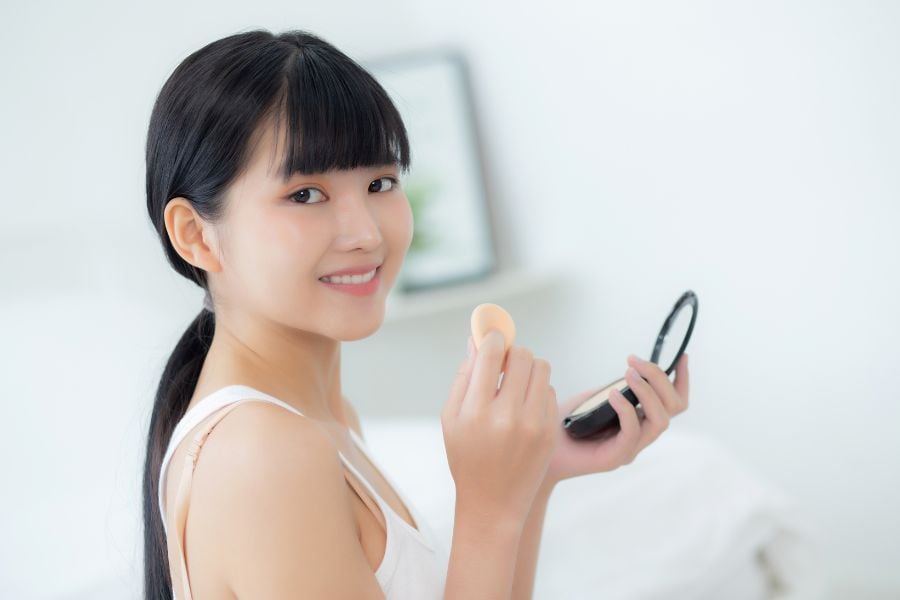 dung-kem-nen-co-can-dung-phan-phu-khong__2__05accd32e0e24138b2103166cd93c102 Người phụ nữ cầm gương trang điểm và bông phấn, mỉm cười nhẹ nhàng trong không gian sáng sủa.