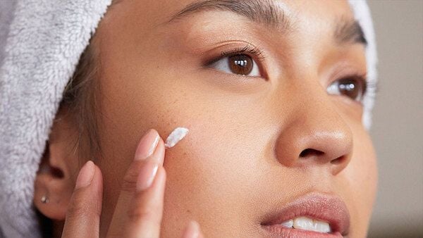 Người phụ nữ đội khăn đang thoa kem dưỡng lên má, nhìn xa xăm.