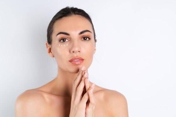 Người phụ nữ với tóc buộc gọn, thoa kem dưỡng dưới mắt, chắp tay trước ngực, nền trắng.