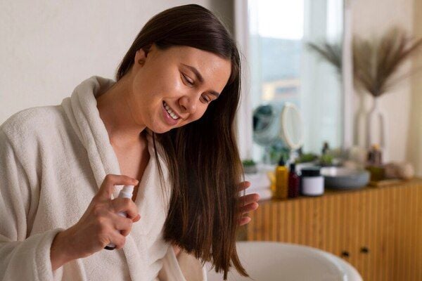Người mặc áo choàng tắm đang xịt dưỡng lên tóc trong phòng có ánh sáng tự nhiên và đồ trang trí đơn giản.