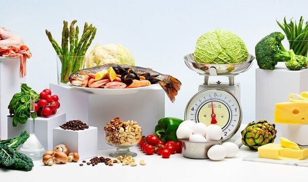 Hỗn hợp thực phẩm gồm rau củ, thịt, cá, trứng, hạt, và phô mai được sắp xếp xung quanh một cái cân.