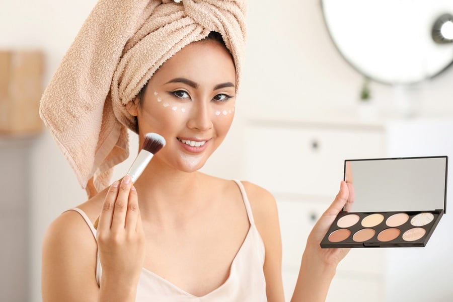 Người cầm bảng phấn trang điểm và cọ, quấn khăn trên đầu, đang chuẩn bị trang điểm.