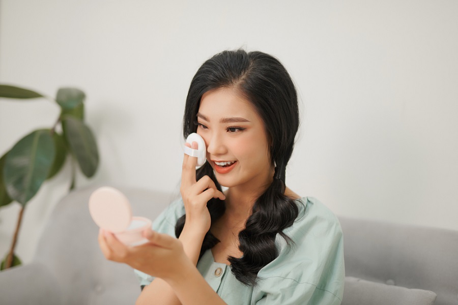 Người phụ nữ đang trang điểm, sử dụng gương cầm tay và bông phấn, ngồi trên ghế sofa, cạnh cây xanh.