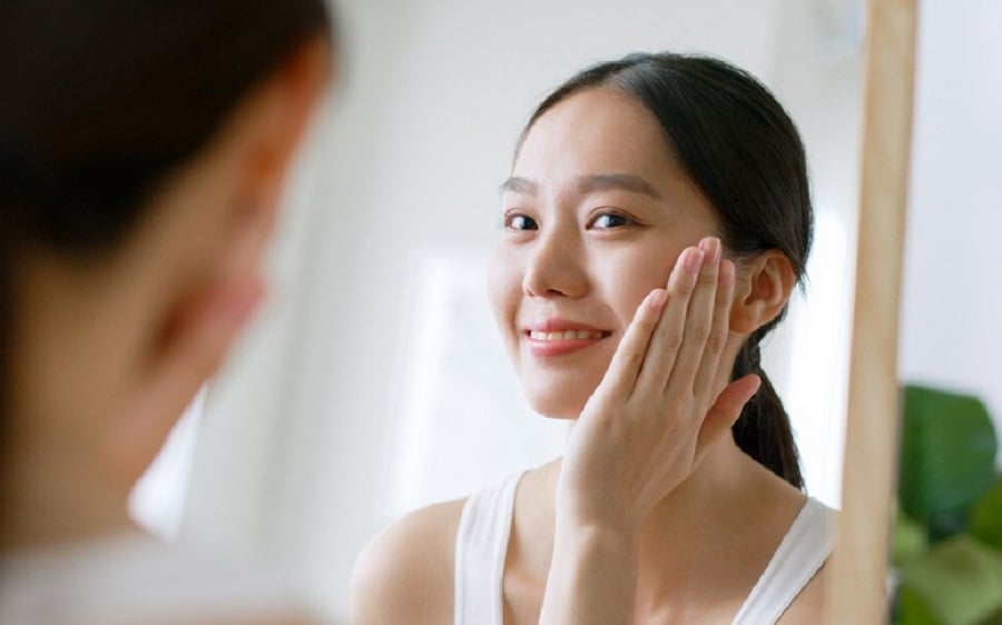 Người phụ nữ cười tươi, chạm tay lên má khi đứng trước gương trong không gian sáng sủa.