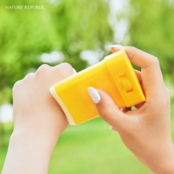 Tay cầm hộp kem chống nắng màu vàng, chuẩn bị thoa lên da, nền xanh mờ của cây cối phía sau.