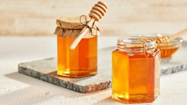 Hai lọ mật ong đặt trên mặt bàn, một lọ có nắp giấy buộc dây và que gỗ khuấy mật.