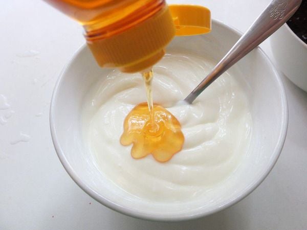 Mật ong đang được rót vào bát sữa chua trắng, có thìa kim loại bên trong.