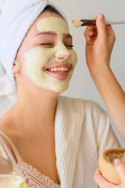 Người phụ nữ đang cười, được thoa mặt nạ dưỡng da trên mặt, quấn khăn trên đầu.