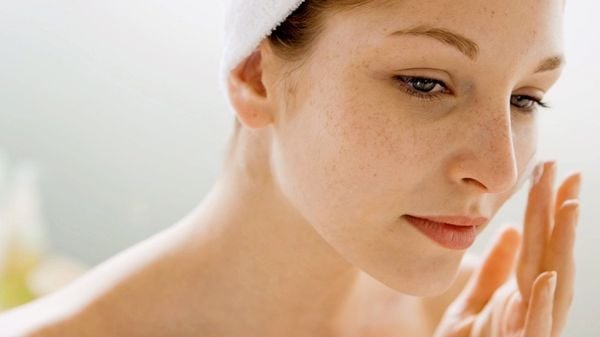 Người phụ nữ với khăn trên đầu đang thoa kem dưỡng lên mặt, nền mờ nhạt phía sau.