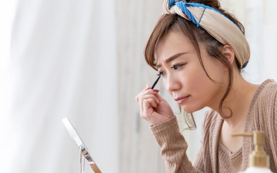 Người phụ nữ đang trang điểm, sử dụng bút kẻ mắt trước gương trong một căn phòng sáng sủa.