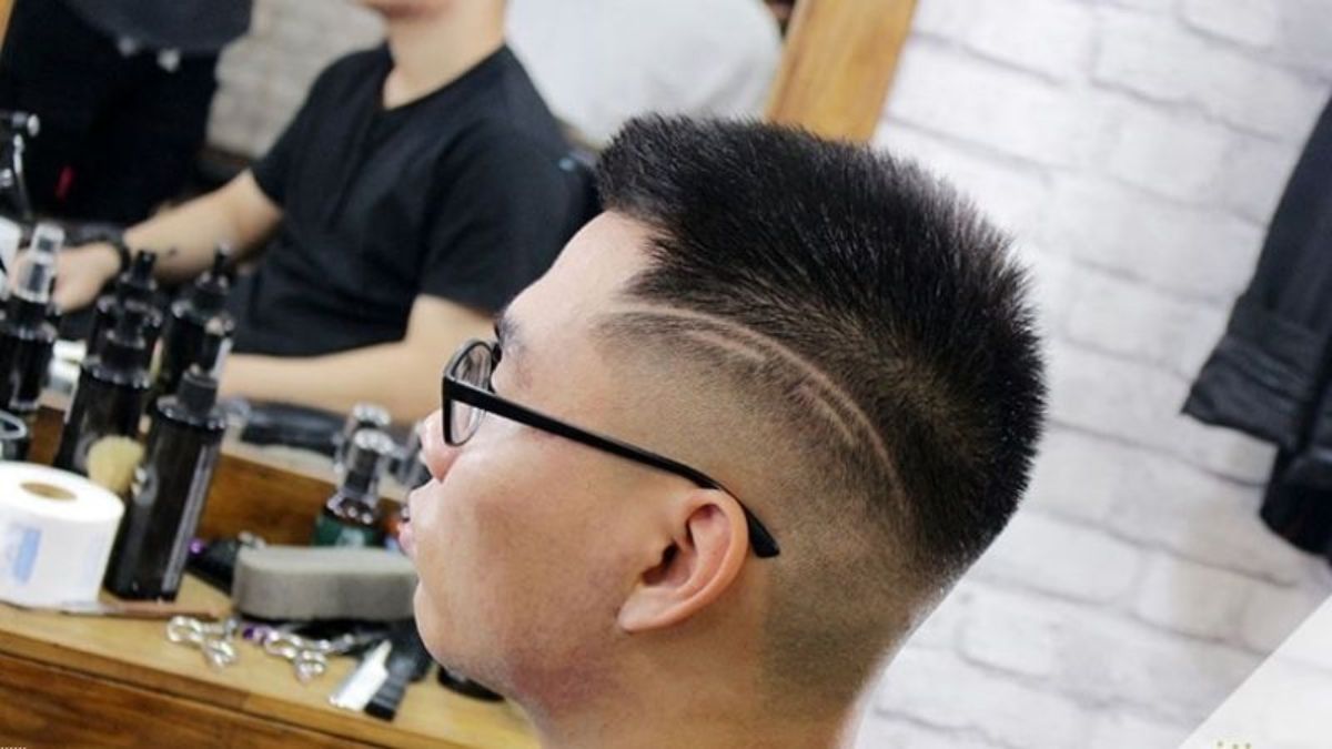 Người đàn ông đeo kính ngồi trong tiệm cắt tóc, mái tóc cắt ngắn với đường tạo kiểu bên trái đầu.