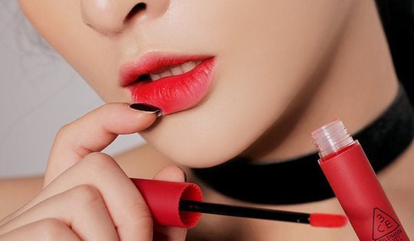 Người mẫu tô son đỏ từ cây cọ. Môi đỏ nổi bật, tay cầm hộp son, nền trắng đơn giản.