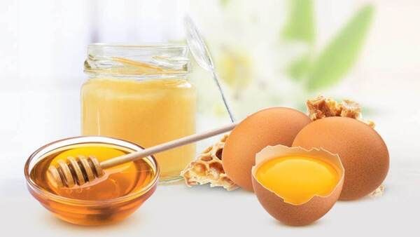 Hũ mật ong, bát mật ong, tổ ong và hai quả trứng, một quả đã tách vỏ, trên nền mờ.